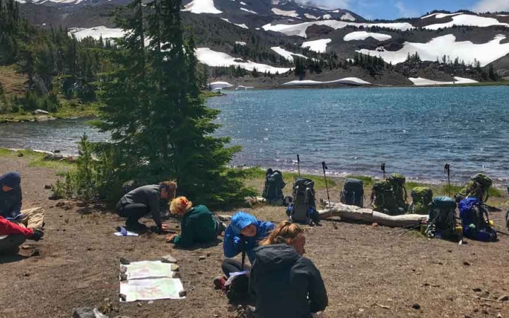 a group of backpacking students rest beside an alpine lake with snowy mountains in the background